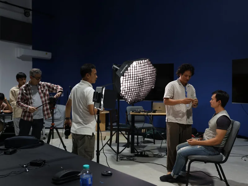 A small crew films an interview subject seated under a softbox, one operator watching the monitor rig while another adjusts the light