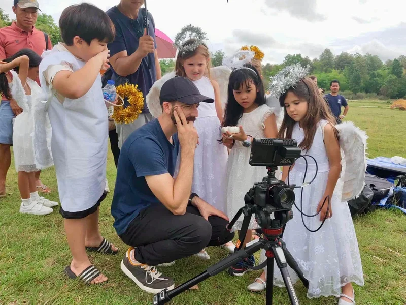 Crouched beside the camera monitor, the director checks a take with a field full of children in angel costumes waiting behind him