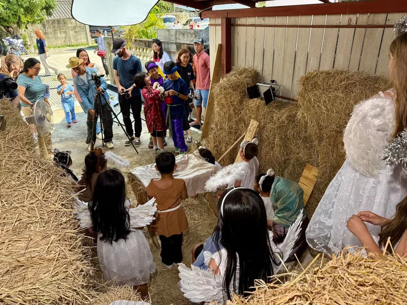 A nativity scene built from stacked hay bales under a carport, child actors in costume gathered around a manger while the crew films from the doorway