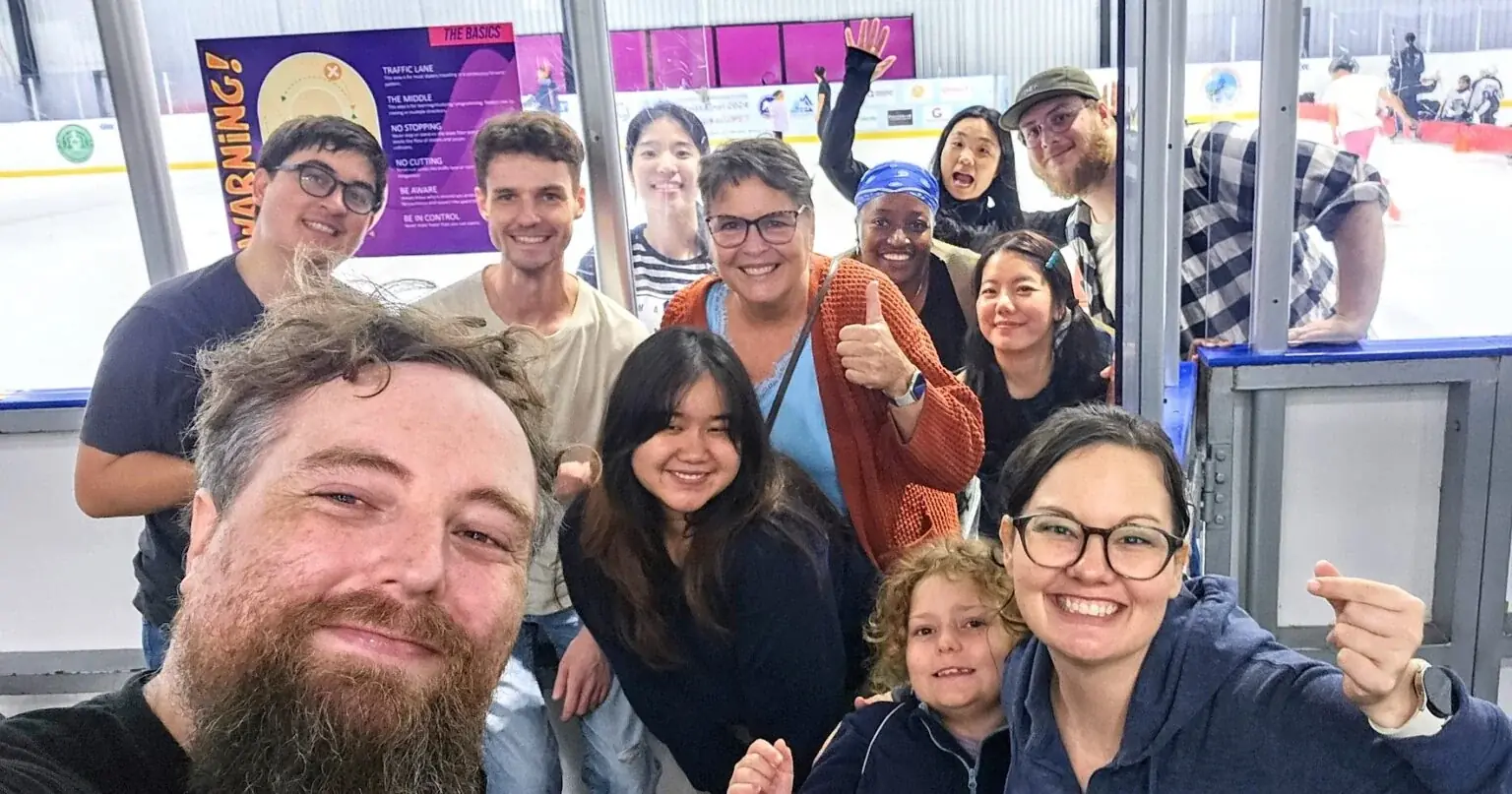 A dozen team members crowd into a selfie at an ice rink, laughing, with skaters on the ice behind them