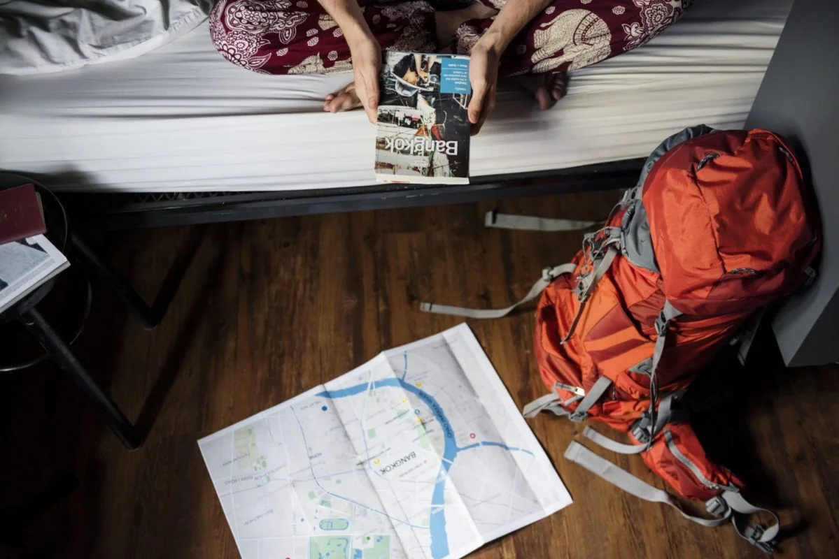 Seen from above, someone sits cross legged on a hostel bed reading a Bangkok guidebook, a packed red rucksack and an open city map on the floor