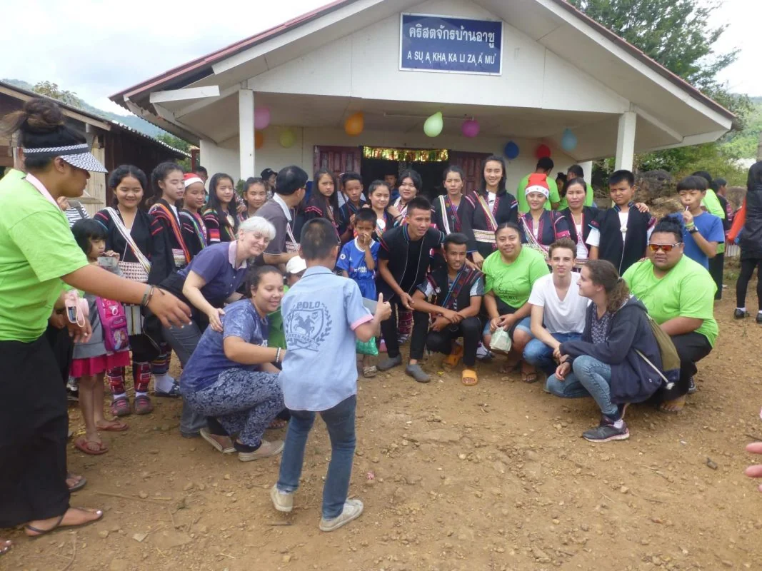 A large group of children, teenagers and adults gathered outside a small village church decorated with balloons, some wearing traditional hill tribe dress