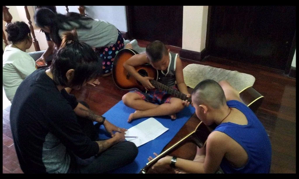 Adults and children sit together on a mat on a wooden floor, two boys learning guitar while a woman writes out chords