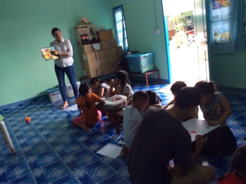 Children sit in groups on a blue tiled floor working on paper while an adult stands at the front holding up a picture book